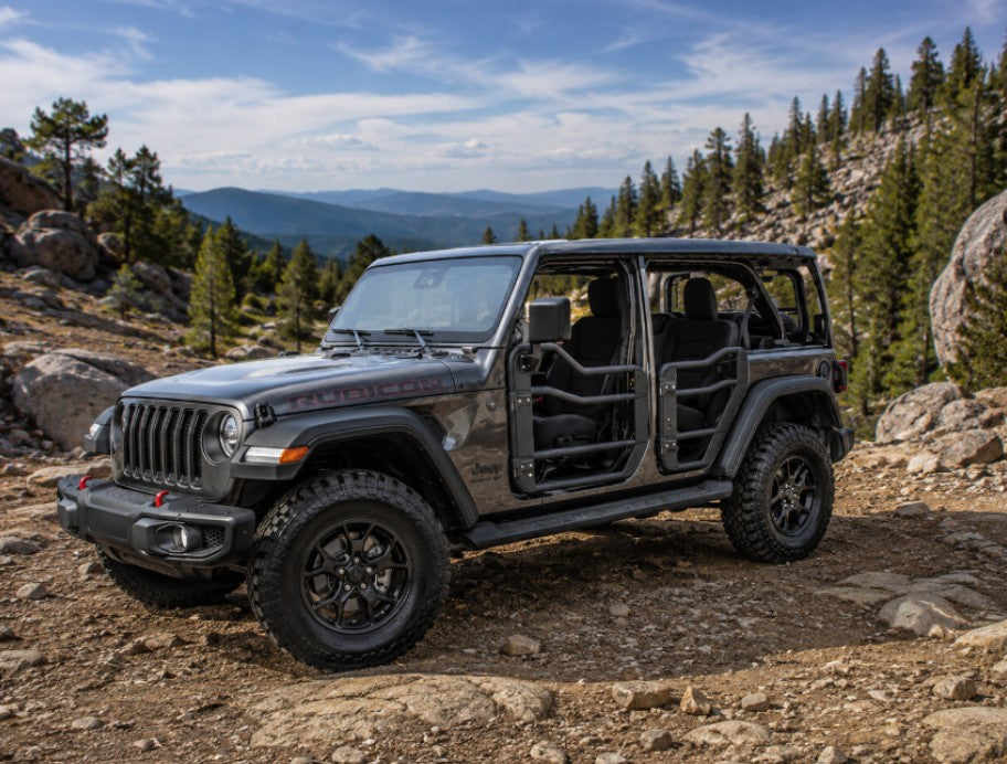 A dark gray Jeep Wrangler with Open tube doors parked on a rocky mountain trail with trees and hills in the background.