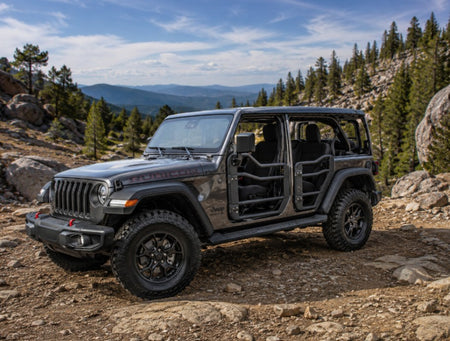A dark gray Jeep Wrangler with Open tube doors parked on a rocky mountain trail with trees and hills in the background.